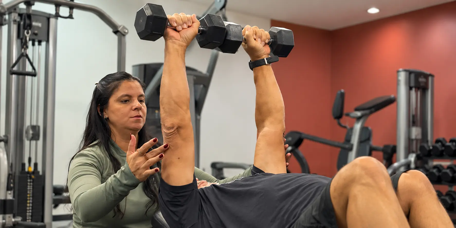 Profissional acompanha de perto um aluno durante exercício com halteres na Everylife